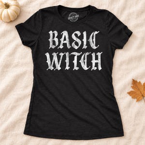 May include: A black short-sleeved t-shirt featuring the text "BASIC WITCH" printed in a distressed white font across the chest. The shirt has a crew neckline and a label inside the collar that reads "CRAZY DOG" with additional small text.
