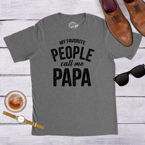 May include: A heather gray short-sleeve t-shirt featuring the black text "MY FAVORITE PEOPLE call me PAPA" printed on the front. The shirt is displayed alongside a pair of brown leather dress shoes, black sunglasses, a wristwatch, and a glass of amber liquid.