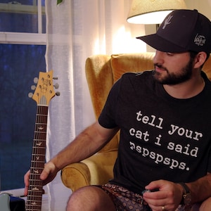 Puede incluir: Una persona con una camiseta negra con el texto "tell your cat i said rspspsps." La persona está sentada en una silla y sostiene una guitarra eléctrica azul.