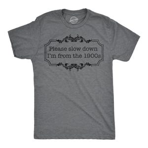 Może przedstawiać: Szary t-shirt z czarnym nadrukiem, na którym widnieje napis "Proszę zwolnić, jestem z lat 1900" w ozdobnej ramce.