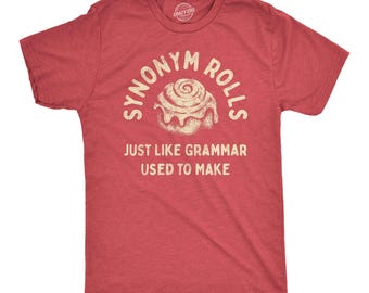 May include: A heathered red short-sleeve t-shirt featuring a graphic of a cinnamon roll in the center. Above the roll is the text "SYNONYM ROLLS" and below it is the text "JUST LIKE GRAMMAR USED TO MAKE" in a distressed, off-white, sans-serif font.