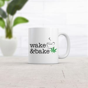 May include: A white ceramic mug featuring the black text wake & bake, a small black line drawing of a steaming coffee cup, and a green cannabis leaf illustration. The mug has a standard handle and a smooth, glossy finish on the exterior surface.