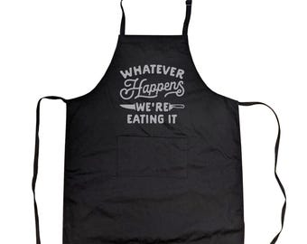 May include: A black apron featuring a central pocket and the light grey text "WHATEVER Happens WE'RE EATING IT" printed on the chest. The design includes a graphic of a kitchen knife positioned between the words "Happens" and "WE'RE".