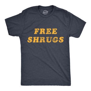 May include: A dark charcoal grey short-sleeved t-shirt featuring the text FREE SHRUGS printed in a distressed, bold yellow font across the chest. The garment has a crew neckline and a standard fit, with a small brand logo visible inside the collar.
