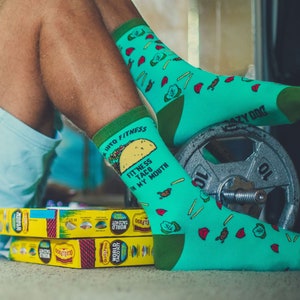 May include: A pair of green and blue socks with a taco graphic and the text "Fitness Taco in My Mouth". The socks are worn on a person's feet, with one sock resting on a weight.
