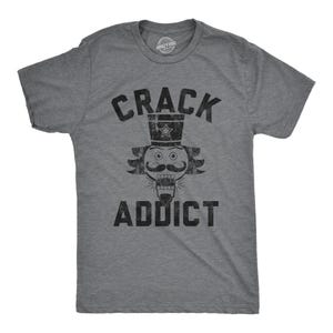 May include: A heather grey short-sleeve t-shirt featuring a black distressed graphic of a nutcracker face wearing a top hat. The text CRACK ADDICT is printed in a bold, distressed black font above and below the nutcracker illustration.