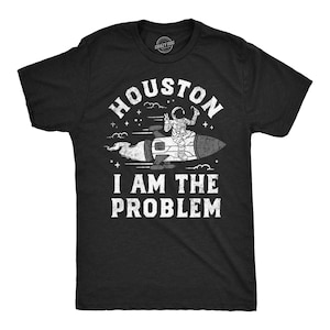 Può includere: Una t-shirt nera a maniche corte con una grafica bianca dall'aspetto vissuto che raffigura un astronauta a cavallo di un razzo. Il testo recita HOUSTON I AM THE PROBLEM. Il design include stelle e nuvole, con un piccolo logo circolare sul collo.