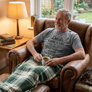 May include: A heather gray short-sleeved t-shirt featuring black text that reads Dads Know A Lot, Grandpas Know Everything with a small anchor graphic in the center. The shirt is worn by an older individual holding a beige ceramic mug.