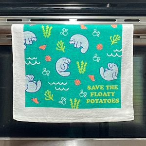 Puede incluir: Una toalla de cocina blanca con fondo verde y un patrón repetido de manatíes, algas marinas y conchas. El texto "SAVE THE FLOATY POTATOES" está impreso en amarillo en la toalla.