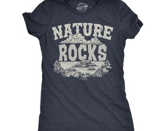 May include: A dark navy blue short-sleeve t-shirt featuring a graphic print in light gray. The design includes the text NATURE ROCKS in a bold font, positioned above an illustration of clouds, a body of water, and several rocky formations.