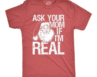 May include: A red short-sleeved t-shirt featuring a white graphic of Santa Claus wearing a hat. The text printed in white reads, ASK YOUR MOM IF I'M REAL. The design has a distressed, weathered appearance on the fabric of the shirt.
