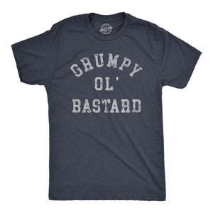 May include: A dark navy blue short-sleeved t-shirt featuring the distressed white text "GRUMPY OL' BASTARD" printed across the chest in a bold, collegiate-style font. The shirt has a crew neckline and a standard fit, with a visible brand logo inside.