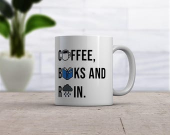 May include: A white ceramic mug featuring black text that reads COFFEE, BOOKS AND RAIN. The text includes a small icon of a coffee mug, an open blue book, and a grey rain cloud with blue raindrops replacing the letters O and A.