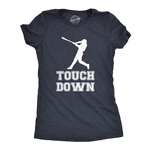 Könnte beinhalten: Ein dunkelblaues T-Shirt mit kurzen Ärmeln und einer weißen Grafik eines Baseballspielers, der einen Schläger schwingt. Unter der Grafik ist der Text "TOUCH DOWN" in einer fetten, weißen Blockschrift gedruckt. Das T-Shirt hat einen Rundhalsausschnitt.