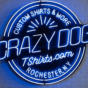 May include: A neon sign for Crazy Dog TShirts & More, featuring a circular design with the text "Custom Shirts & More" and "Crazy Dog" in a bold font. The sign also includes the website address and the city, Rochester, NY.