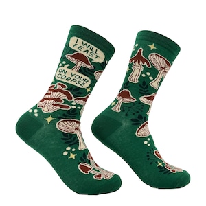 Peut inclure: Une paire de chaussettes montantes vert foncé avec un motif répétitif de champignons marron et crème, de feuillage vert foncé et de petites étoiles jaunes. Une chaussette porte une bulle avec le texte I WILL FEAST ON YOUR CORPSE.