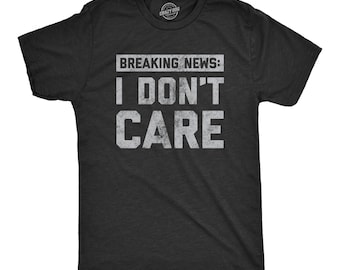 Puede incluir: Una camiseta gris carbón de manga corta con el texto blanco desgastado BREAKING NEWS: I DON'T CARE impreso en el pecho. La camiseta tiene cuello redondo y un corte estándar, con un pequeño logotipo de marca circular visible en el cuello.