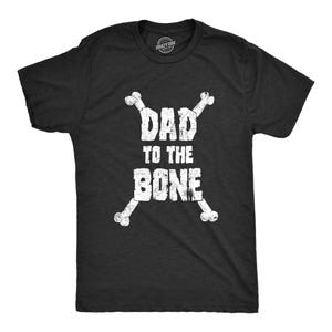 May include: A black short-sleeved t-shirt featuring a white distressed graphic print. The design displays the text DAD TO THE BONE in a block font, positioned between two crossed white bones that form an X shape behind the lettering.