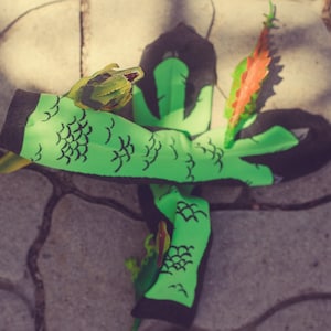 May include: A pair of bright green socks featuring a black scale pattern and black cuffs. The socks are adorned with small, attached plastic dinosaur figures in shades of green and orange, positioned along the fabric surface.