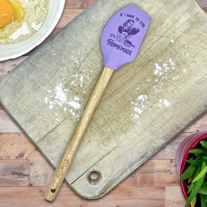 May include: A lavender silicone spatula with a wooden handle. The spatula head features a dark purple graphic of a person cooking and the text "IF I HAVE TO STIR IT'S HOMEMADE". The handle is light brown wood with a small hole at the bottom end.