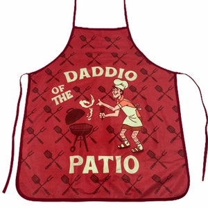 Op de afbeelding: Rode schort met een witte afbeelding van een man die barbecuet en de tekst "Daddio of the Patio". De schort heeft een herhalend patroon van gekruiste grillvorken.