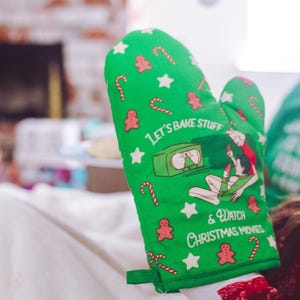May include: A green oven mitt featuring a pattern of red gingerbread men, white stars, and red and white candy canes. The center graphic shows a vintage television set and the text "LET'S BAKE STUFF & WATCH CHRISTMAS MOVIES" in white.