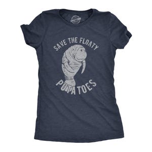 May include: A dark navy blue short-sleeve t-shirt featuring a distressed light gray graphic of a manatee in the center. The text "SAVE THE FLOATY POTATOES" is printed in a distressed light gray font arched above and below the manatee illustration.