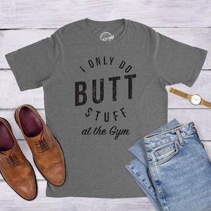 May include: A charcoal gray short-sleeve t-shirt with the black text I ONLY DO BUTT STUFF at the gym printed on the front. The shirt is displayed alongside a pair of brown leather dress shoes, a pair of blue denim jeans, and a gold wristwatch.