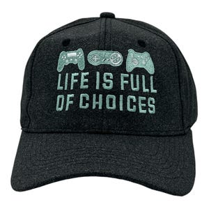 May include: A dark charcoal gray baseball cap featuring light mint green embroidery. The design includes three different video game controllers above the text "LIFE IS FULL OF CHOICES" stitched in a bold, sans-serif font on the front panel.