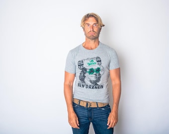 May include: A heather gray short-sleeve t-shirt featuring a graphic of Benjamin Franklin wearing a white cap with a green shamrock and green shamrock-shaped glasses. The text "BEN DRANKIN" is printed in black below the graphic illustration.