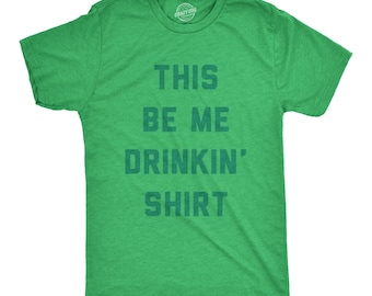 May include: A heather green short-sleeve t-shirt featuring the dark green text THIS BE ME DRINKIN' SHIRT printed in a bold, block-style font across the chest. The garment has a crew neckline and a standard fit, with a visible brand logo on the inner neck.