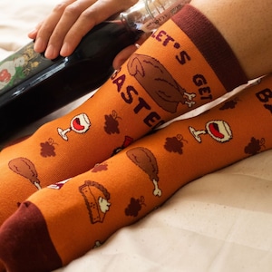 May include: A pair of orange socks with a Thanksgiving theme. The socks feature images of turkeys, wine glasses, and pies. The text on the socks reads "Let's Get Basted".