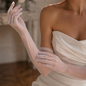 Peut inclure: Une paire de longs gants opéra blancs et transparents, confectionnés dans un tissu léger et translucide. Les gants remontent au-dessus des coudes, présentant une silhouette lisse et ajustée couvrant les mains et les bras, portés avec une robe blanche sans bretelles.
