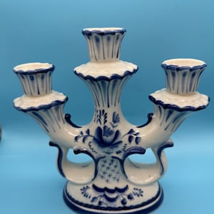 Könnte beinhalten: Ein weißer Keramik-Kerzenständer mit drei Kerzenhaltern mit gewelltem Rand. Das Stück ist mit handgemalten blauen Blumenmustern und feinen Linien verziert, darunter ein zentrales Blumenmotiv und ein Gittermuster auf dem Sockel.