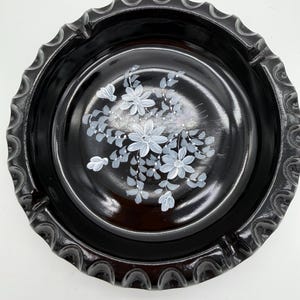 May include: A round, black ashtray with a scalloped edge. The interior features a hand-painted floral design in white and gray. The ashtray is made of a glossy material.