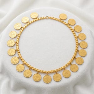 Puede incluir: Un collar dorado con un diseño de cadena y múltiples colgantes de monedas. Las monedas son redondas y tienen grabados detallados. El collar se muestra sobre una superficie blanca.