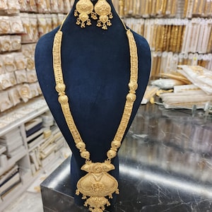 Puede incluir: Un conjunto de collar y pendientes en tono dorado con intrincados patrones de filigrana. El collar largo incluye eslabones rectangulares texturizados, cuentas esféricas y un gran colgante ornamentado con elementos colgantes a juego.