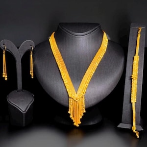Puede incluir: Un conjunto de joyas de color dorado exhibido en soportes negros. El conjunto incluye un collar con diseño en forma de V y flecos de cuentas, aretes a juego y una pulsera. Las joyas se presentan sobre un fondo oscuro.
