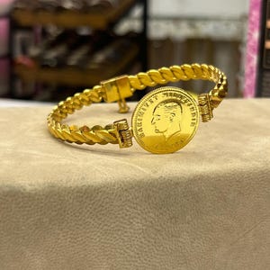 May include: A gold-colored bracelet featuring a braided chain design and a central circular coin charm. The coin displays a profile portrait of a person facing left, with the text "HAKIMIYET" clearly visible along the edge of the coin surface.