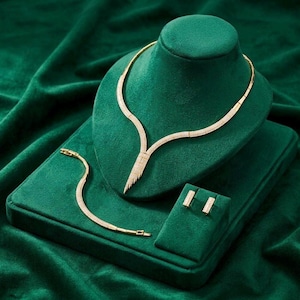 Puede incluir: Un conjunto de joyería en tono dorado que incluye un collar en forma de V con un colgante puntiagudo y texturizado, una pulsera fina a juego con cierre y un par de pendientes rectangulares pequeños, adornados con pequeñas piedras claras.
