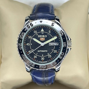 May include: A Seiko automatic wristwatch with a black dial, luminous hands, and a date display. The watch has a silver bezel with minute markers and a blue leather strap. The watch face displays the text "SEIKO" and "AUTOMATIC".