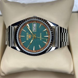 Puede incluir: Un reloj de pulsera autom&aacute;tico Seiko 5 plateado con esfera verde y correa plateada. La esfera muestra el d&iacute;a y la fecha, con agujas e indicadores dorados. El reloj se presenta sobre un coj&iacute;n color crema.