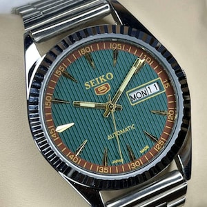 Puede incluir: Un reloj de pulsera automático Seiko con esfera verde y agujas e indicadores dorados. El reloj tiene una correa de acero inoxidable y una pantalla de fecha que muestra "LUN 11". La esfera del reloj tiene un patrón texturizado y un anillo exterior marrón.