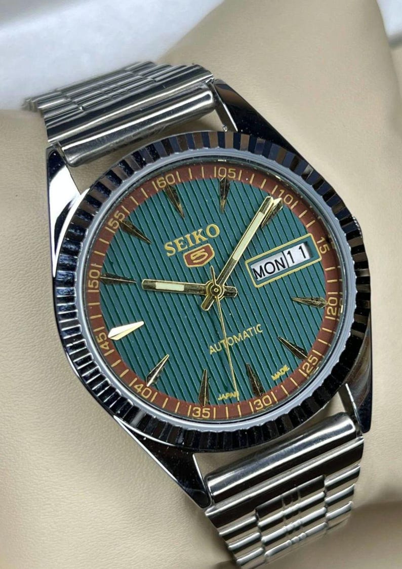 Puede incluir: Un reloj de pulsera autom&aacute;tico Seiko con esfera verde y agujas e indicadores dorados. El reloj tiene una correa de acero inoxidable y una pantalla de fecha que muestra "LUN 11". La esfera del reloj tiene un patr&oacute;n texturizado y un anillo exterior marr&oacute;n.