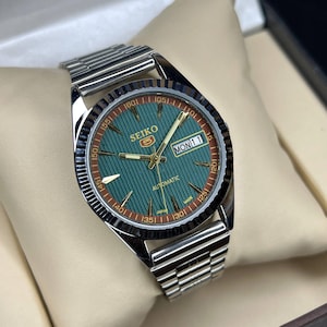 Puede incluir: Un reloj autom&aacute;tico Seiko plateado con esfera verde y correa plateada. La esfera muestra el d&iacute;a y la fecha, con agujas y marcadores dorados. El reloj se presenta sobre un coj&iacute;n color crema.