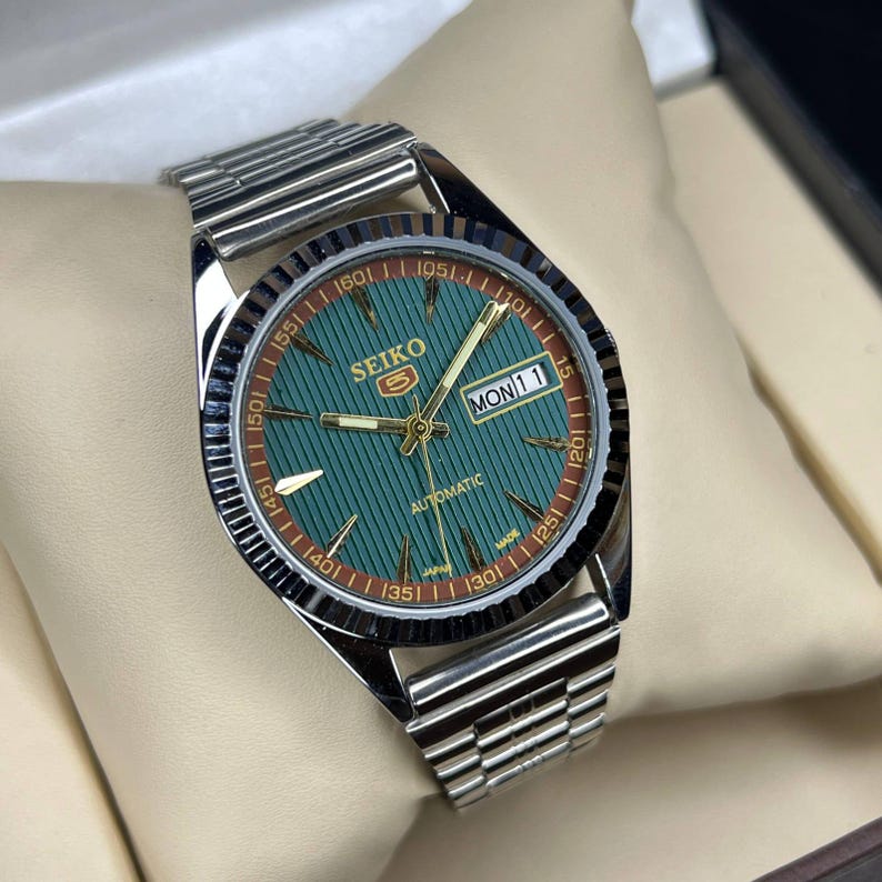 Puede incluir: Un reloj autom&aacute;tico Seiko plateado con esfera verde y correa plateada. La esfera muestra el d&iacute;a y la fecha, con agujas y marcadores dorados. El reloj se presenta sobre un coj&iacute;n color crema.