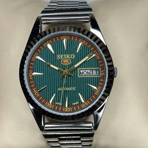 Puede incluir: Un reloj autom&aacute;tico Seiko 5 plateado con esfera verde y visualizaci&oacute;n de d&iacute;a y fecha. El reloj presenta agujas e indicadores dorados y una correa de acero inoxidable. La esfera tiene un patr&oacute;n de rayas verticales y el d&iacute;a se muestra como "MON 11".