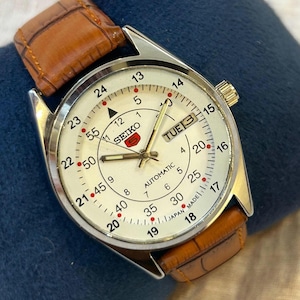 Op de afbeelding: Een vintage Seiko 5 automatische polshorloge met een witte wijzerplaat, zilveren kast en bruine leren band. De wijzerplaat toont de dag en datum, met een 24-uurs schaal. Het horloge is gemarkeerd met "Japan Made".