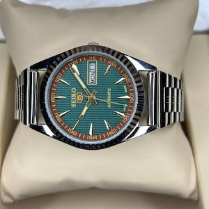 Puede incluir: Un reloj autom&aacute;tico Seiko 5 plateado con esfera verde y correa plateada. La esfera muestra el d&iacute;a y la fecha, con agujas e indicadores dorados. El reloj se presenta sobre un coj&iacute;n color crema.