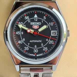 Puede incluir: Reloj automático Seiko de acero inoxidable con esfera negra. La esfera presenta números blancos, una ventana de fecha y agujas luminosas. La correa del reloj también es de acero inoxidable.